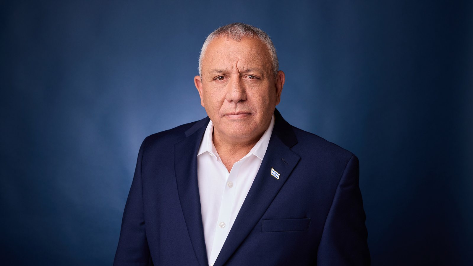 Gadi Eisenkot, in a navy suit, white open-collar shirt and Israeli flag lapel pin, stands in front of a dark blue studio backdrop