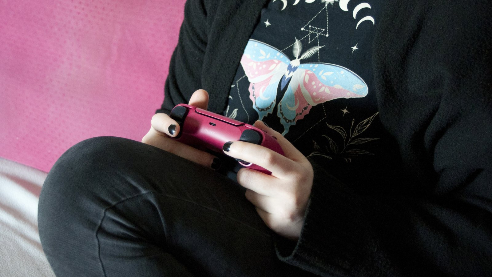 A person in a black butterfly T-shirt sits on a pink couch holding a pink PlayStation 5 DualSense controller with both hands
