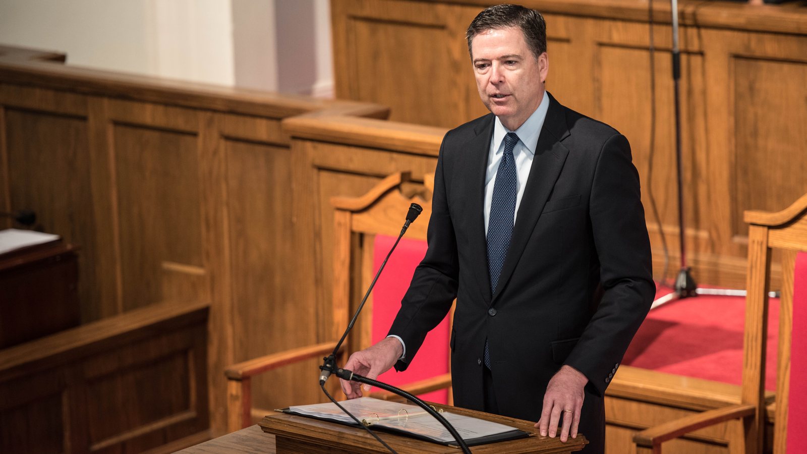 James Comey in a dark suit and blue tie speaking from a wooden lectern with a microphone, red chairs and wood paneling behind him