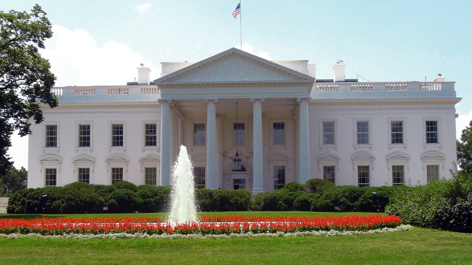 The north facade of the White House in Washington, with its columned portico, and a fountain on the North Lawn in front, on a clear day