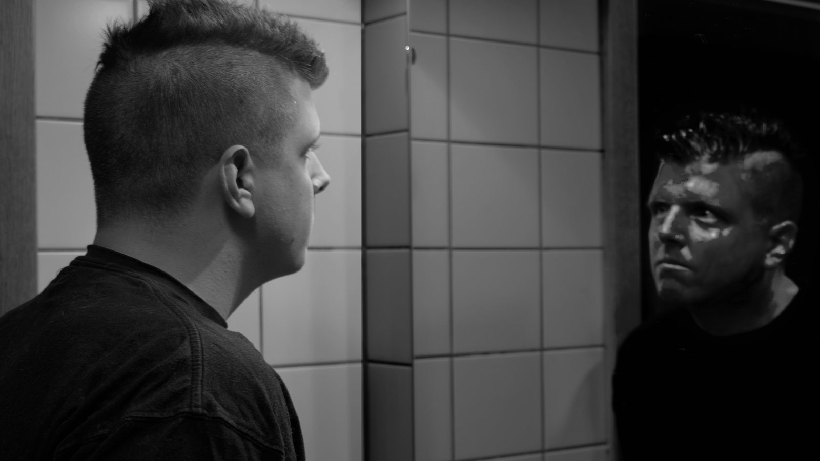 A man with a short mohawk haircut studies his reflection in a bathroom mirror, in black and white