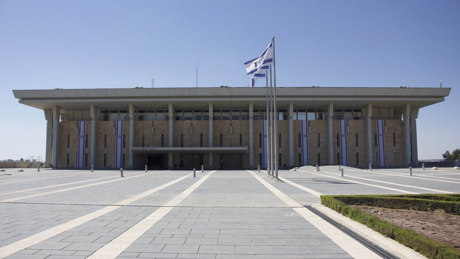 The front of the Knesset building in Jerusalem under a clear blue sky, with Israeli flags on a pole in the plaza and banners on the facade