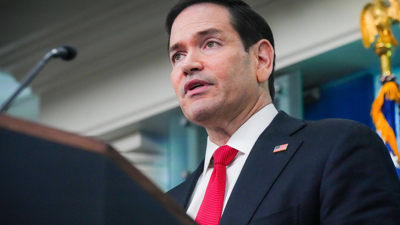 Secretary of State Marco Rubio, in a dark suit and red tie, speaks at the White House briefing room podium