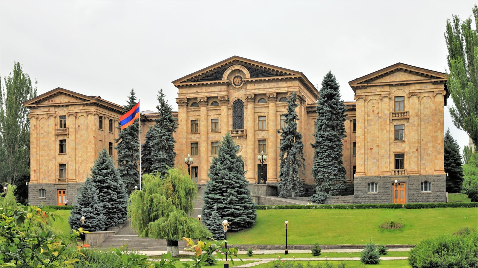 The National Assembly building in Yerevan, Armenia, a long stone facade with columns and a flag, behind fir trees and a lawn