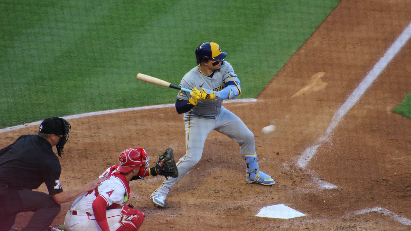 Brewers catcher William Contreras, in a gray road uniform and navy and gold helmet, watches a pitch from the batter's box as an Angels catcher and the umpire crouch behind the plate