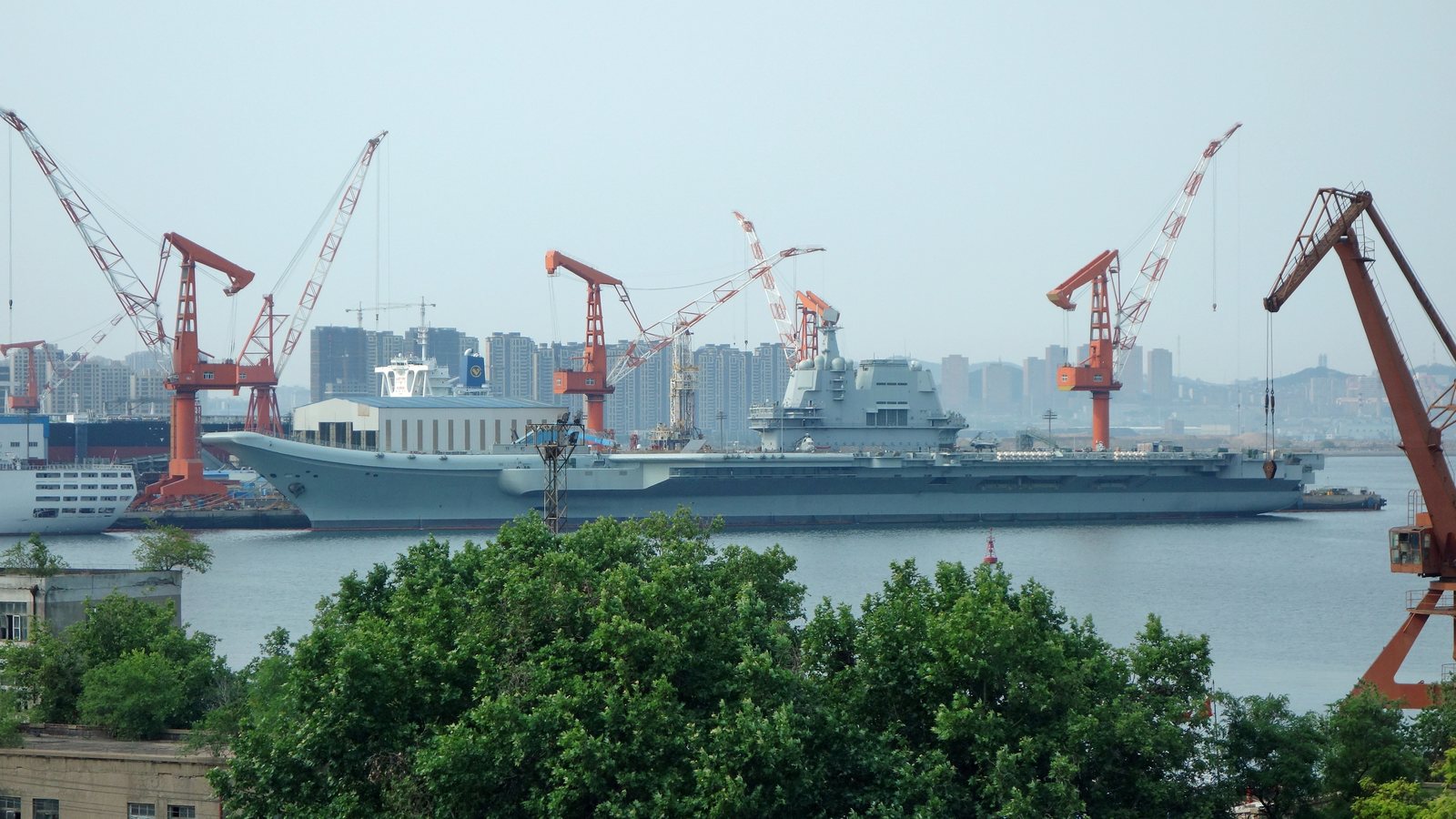 A gray aircraft carrier docked beside orange shipyard cranes, with apartment towers behind it