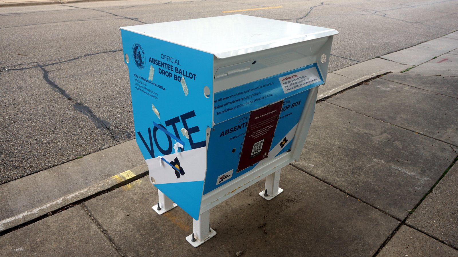 A blue and white official absentee ballot drop box marked Vote on a sidewalk in Madison, Wisconsin