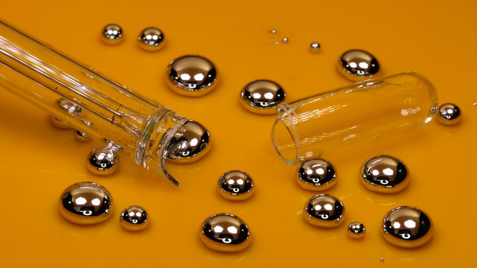 Silvery droplets of liquid mercury and a broken glass thermometer on an orange-gold surface