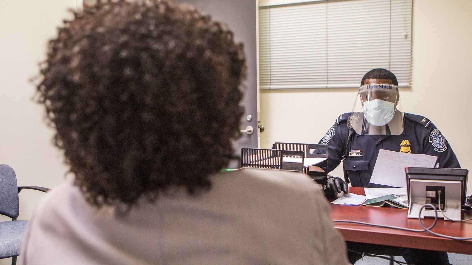 A US Customs and Border Protection officer in a face shield and mask interviews an arriving passenger at a desk at Washington Dulles airport