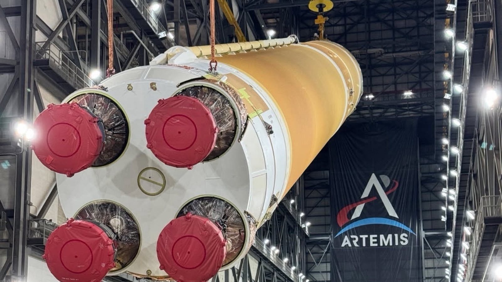The four engine nozzles of NASA's orange Artemis III SLS core stage, covered in red caps, as cranes lift the stage inside the Vehicle Assembly Building under an Artemis banner