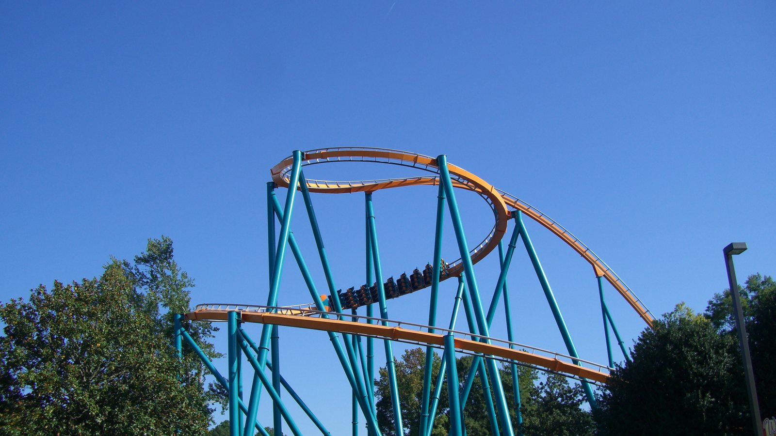 The orange track and teal supports of the Goliath roller coaster at Six Flags Over Georgia, with a train passing through a helix against a blue sky