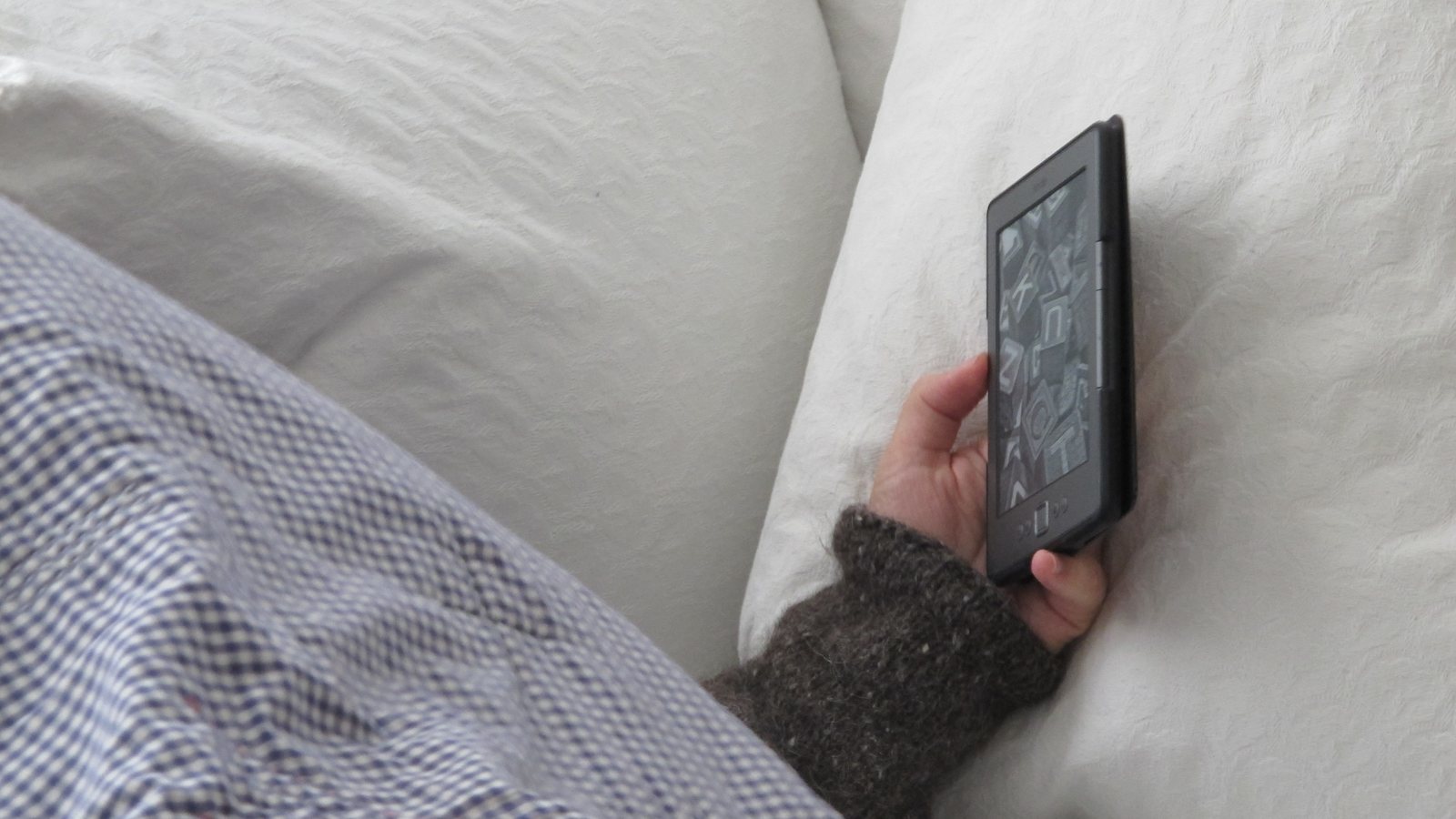 A person napping in bed under a checked blanket, one hand holding an e-reader against a white pillow