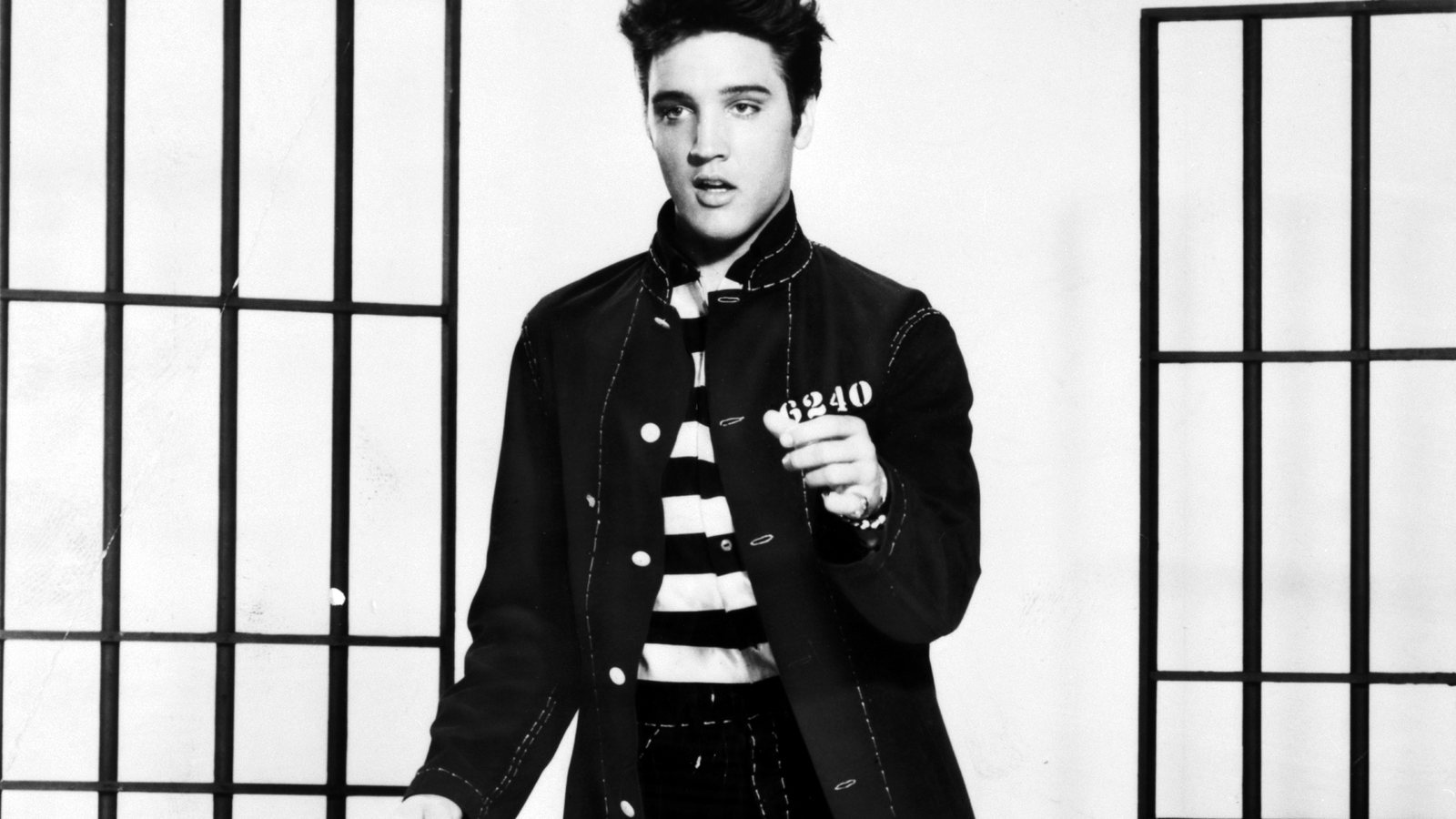 Elvis Presley in a dark jacket over a striped shirt, standing between prop jail bars in a black-and-white 1957 publicity photo for Jailhouse Rock