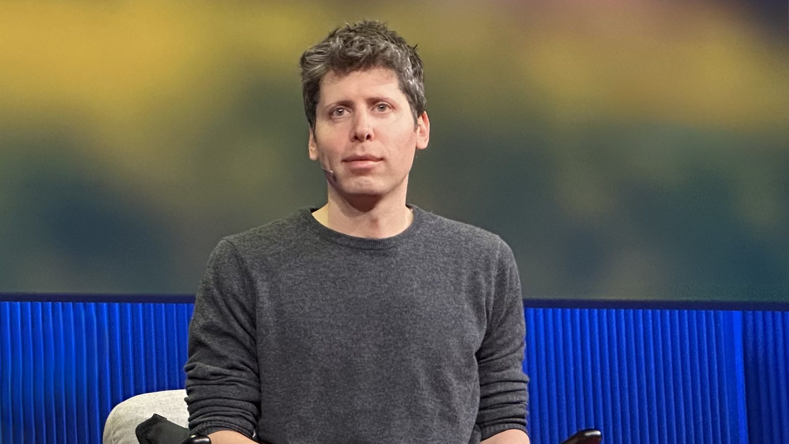 OpenAI chief executive Sam Altman, in a gray sweater, seated on the TED stage in front of a blue and gold backdrop