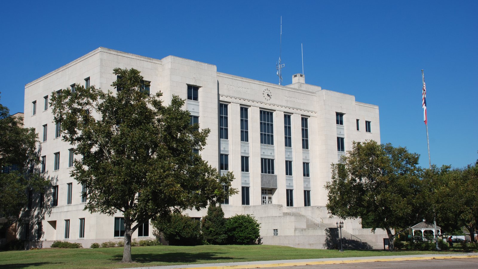 The white stone Brazoria County Courthouse in Angleton, Texas, behind trees and a lawn under a blue sky