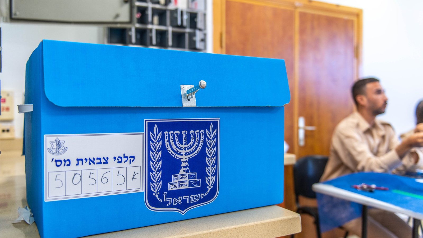 A blue Israeli ballot box with the state emblem and a numbered label on a counter at a military polling station