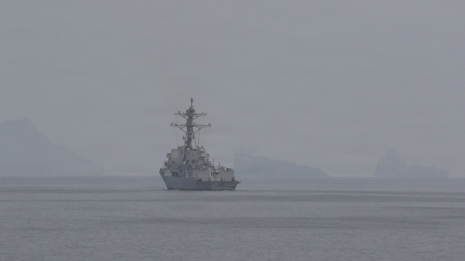 A US Navy guided-missile destroyer on hazy water in the Strait of Hormuz, with a helicopter overhead and rocky islands behind